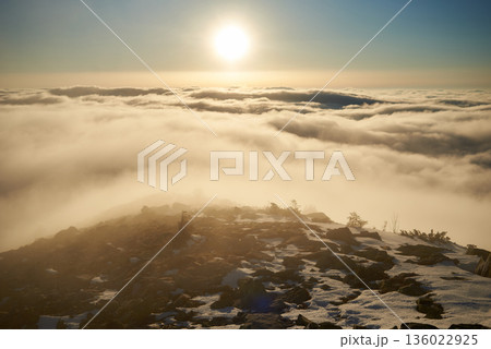 Sunrise over snow-covered mountain peak. Warm golden light of rising sun bathes clouds in soft hues of orange and pink. Snow and scattered rocks in foreground add texture and depth to scene. Sunrise over snow-covered mountain peak. Warm golden light of rising sun bathes clouds in soft hues of orange and pink. Snow and scattered rocks in foreground add texture and depth to scene. 136022925