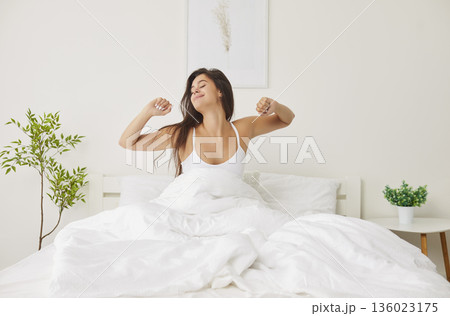 Portrait of a happy smiling young woman waking up in bed stretching her arms in modern bedroom. Portrait of a happy smiling young woman waking up in bed stretching her arms in modern bedroom. 136023175