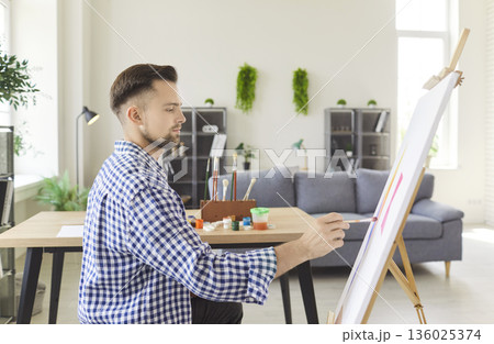 young man artist sitting in front of easel at home workshop, making picture 136025374