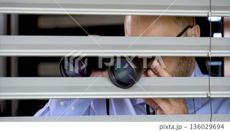 Man looks through blinds with binoculars in an office setting during daylight hours Man looks through blinds with binoculars in an office setting during daylight hours 136029694