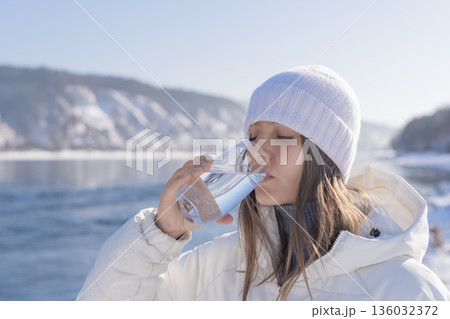 A woman wearing warm clothing drinks fresh water from a glass while standing near a river in winter. Snow covers the ground and mountains are visible in the background. High quality photo 136032372