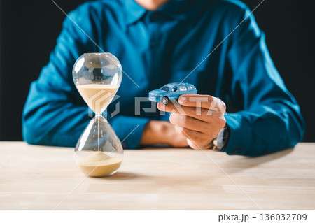 Man hand holds small blue car near hourglass on wood table, sand falling through symbolizes time passing for car loan finance, saving money, and insurance cost Man hand holds small blue car near hourglass on wood table, sand falling through symbolizes time passing for car loan finance, saving money, and insurance cost 136032709