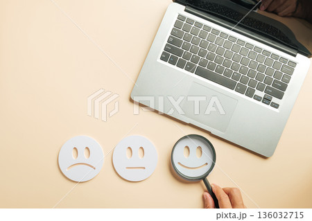 Online customer review feedback shows various satisfaction levels, hand holds magnifying glass choosing happy face near laptop, concept for digital survey and client assessment needs Online customer review feedback shows various satisfaction levels, hand holds magnifying glass choosing happy face near laptop, concept for digital survey and client assessment needs 136032715