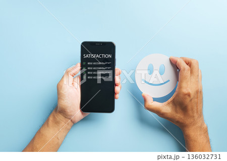 Star customer online review experience via mobile phone, hands holding smartphone with satisfaction survey for service feedback, smiling face cutout showing excellent rating 136032731