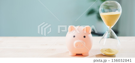 Pink piggy bank money invest dividend income interest fund pig save concept stands beside hourglass on wooden table, representing time management and productivity for wealth growth 136032841