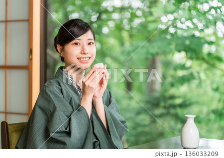 旅館の客室の縁側で日本酒を飲みながらくつろぐ浴衣羽織姿の若いアジア人女性 旅館の客室の縁側で日本酒を飲みながらくつろぐ浴衣羽織姿の若いアジア人女性 136033209