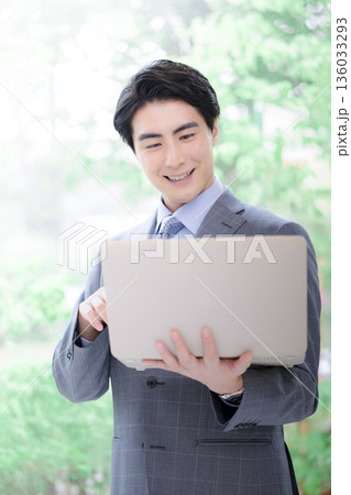 逆光の柔らかい新緑の中でパソコンを持つスーツの男性の笑顔 コンサルタント、リクルートなど転職 逆光の柔らかい新緑の中でパソコンを持つスーツの男性の笑顔 コンサルタント、リクルートなど転職 136033293