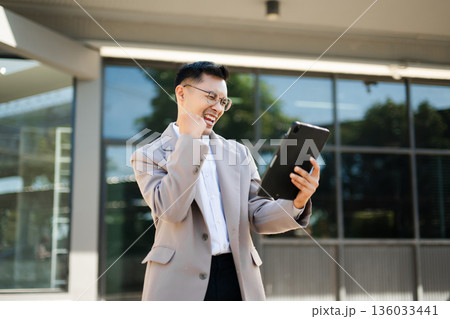 Happy businessman raising fist and cheering with smartphone in hand, celebrating success and victory with excitement and joy outdoors. 136033441