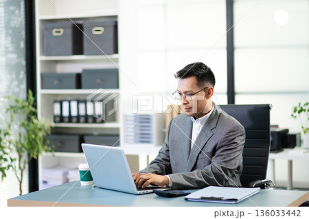 Asian man using laptop and tablet while sitting at her working place. Concentrated at work. Asian man using laptop and tablet while sitting at her working place. Concentrated at work. 136033442