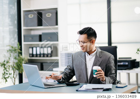A professional businessman working on a laptop while holding a coffee cup in a modern office, representing productivity A professional businessman working on a laptop while holding a coffee cup in a modern office, representing productivity 136033489