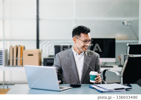 A professional businessman working on a laptop while holding a coffee cup in a modern office, representing productivity 136033490