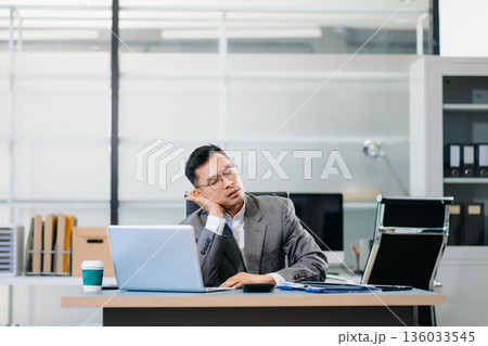 Exhausted businessman sitting at desk with laptop, stressed by workload, deadlines, and corporate pressure in modern office environment. Exhausted businessman sitting at desk with laptop, stressed by workload, deadlines, and corporate pressure in modern office environment. 136033545