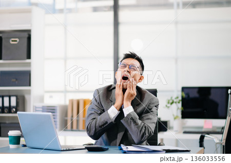 Tired businessman sitting at office desk with laptop, feeling stressed and overworked under tight deadlines and corporate pressure. Tired businessman sitting at office desk with laptop, feeling stressed and overworked under tight deadlines and corporate pressure. 136033556
