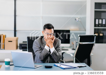 Tired businessman sitting at office desk with laptop, feeling stressed and overworked under tight deadlines 136033559