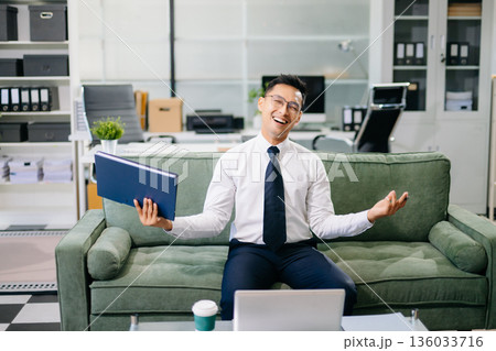 Asian businessman sitting on office sofa using smartphone and laptop, smiling while working with documents in a modern workspace. 136033716
