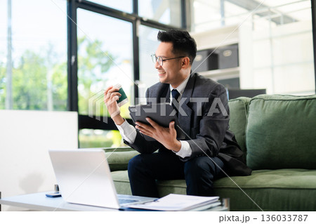 Smiling businessman using laptop and smartphone while working in modern office, digital communication, mobile technology, productivity Smiling businessman using laptop and smartphone while working in modern office, digital communication, mobile technology, productivity 136033787