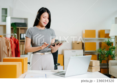 Startup small business SME, Entrepreneur owner woman using smartphone or tablet taking receive and checking online purchase shopping order to preparing pack product box. Startup small business SME, Entrepreneur owner woman using smartphone or tablet taking receive and checking online purchase shopping order to preparing pack product box. 136034320