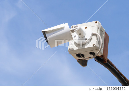 A white CCTV surveillance camera on an outdoor street against a background of blue sky in close-up. Security and surveillance, monitoring of the street situation in a criminal area. High quality photo 136034332