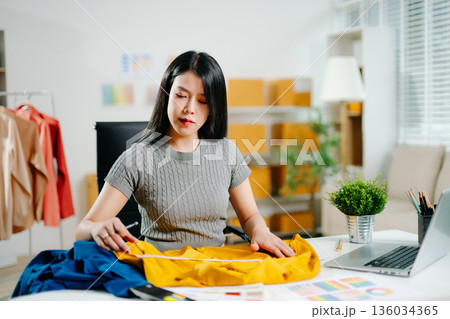 Asian fashion designer measuring fabric at home studio with mannequin and laptop. Creative fashion business, tailor planning clothing design Asian fashion designer measuring fabric at home studio with mannequin and laptop. Creative fashion business, tailor planning clothing design 136034365
