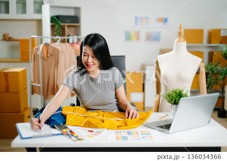 Asian fashion designer measuring fabric at home studio with mannequin and laptop. Creative fashion business, tailor planning clothing design 136034366