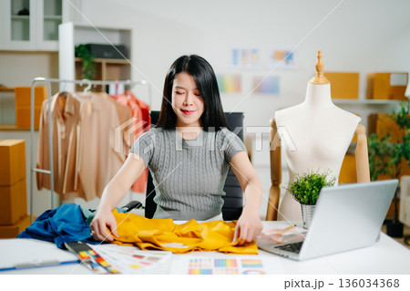 Asian fashion designer measuring fabric at home studio with mannequin and laptop. Creative fashion business, tailor planning clothing design 136034368