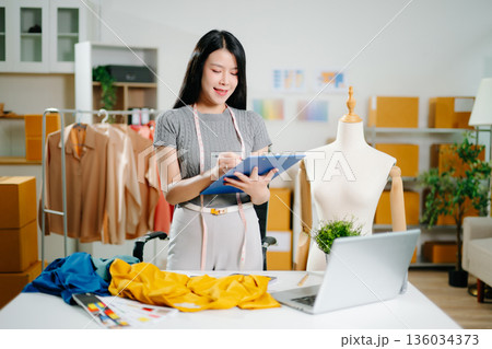 Asian fashion designer working in studio with mannequin, fabric and laptop. Creative fashion business, tailor planning clothing design 136034373