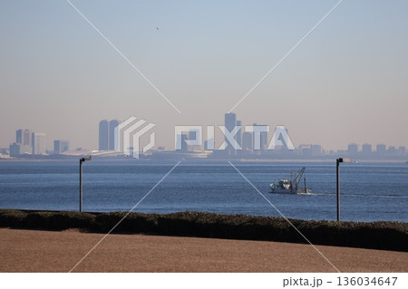 青空と霞む高層ビルの街並みと青い海面の海と漁船と海岸の生け垣と冬枯れの芝生の公園の風景 青空と霞む高層ビルの街並みと青い海面の海と漁船と海岸の生け垣と冬枯れの芝生の公園の風景 136034647