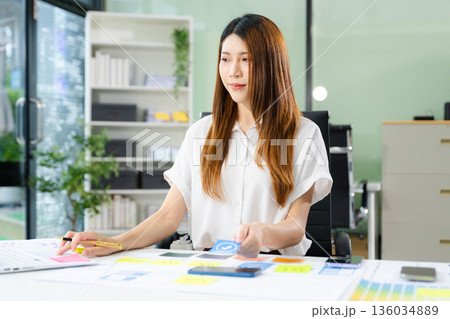 Creative Asian UX UI designer planning mobile app layout with sticky notes, color swatches, and wireframes. 136034889