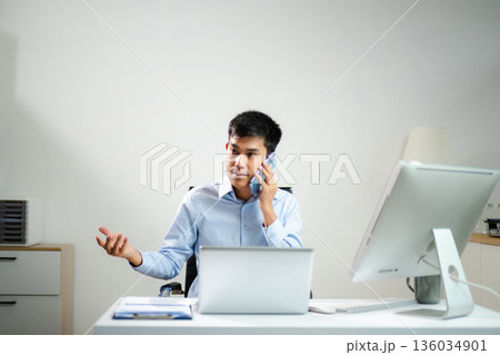 Asian businessman multitasking on phone and monitor at desk. Modern office tech, communication, and career success in action. 136034901