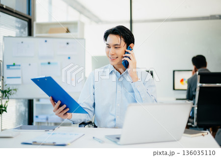 Asian Businessman Talking on the phone and using a laptop with a smile while sitting at office Asian Businessman Talking on the phone and using a laptop with a smile while sitting at office 136035110