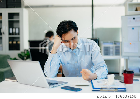 Asian man feeling migraine head strain.Overworked businessman financier while working on laptop and tablet at office. Asian man feeling migraine head strain.Overworked businessman financier while working on laptop and tablet at office. 136035159