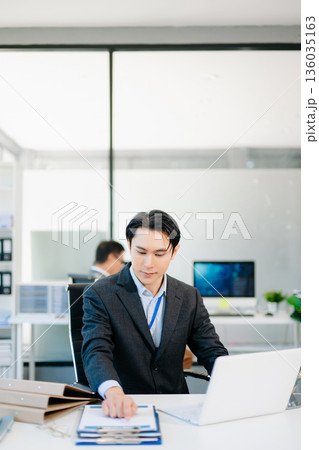 Confident Asian businessman in suit working on laptop and clipboard at bright modern office, symbolizing professionalism, success, and corporate teamwork. 136035163