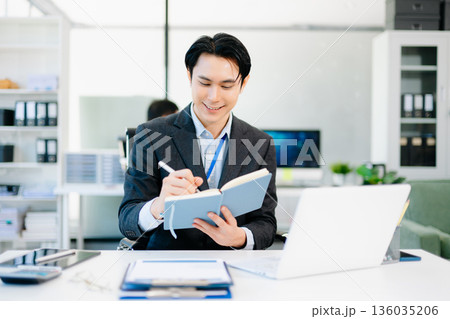 Confident Asian businessman writing notes in notebook while working at bright modern office 136035206