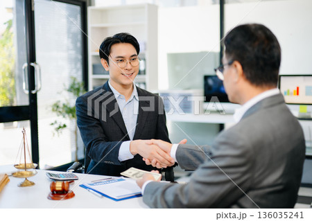 Lawyer and client shaking hands after contract signing with money and gavel on desk. Ideal for business, finance, partnership, or legal themes. Lawyer and client shaking hands after contract signing with money and gavel on desk. Ideal for business, finance, partnership, or legal themes. 136035241