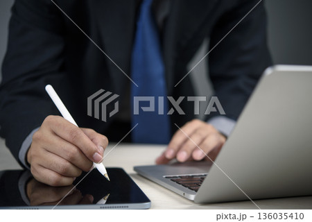 Businessman signing digital document on tablet with stylus while working on laptop, modern technology for business contract and electronic signature concept. 136035410