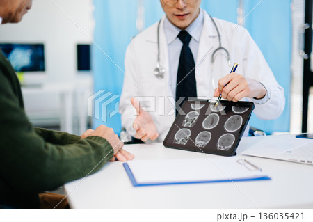 Doctor showing brain MRI results to patient during consultation in modern clinic. Perfect for healthcare, diagnosis, neurology, or medical treatment themes. Doctor showing brain MRI results to patient during consultation in modern clinic. Perfect for healthcare, diagnosis, neurology, or medical treatment themes. 136035421