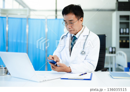 Asian doctor working on laptop and writing notes in modern clinic. Ideal for healthcare, diagnosis, medical research, and professional consultation themes. 136035515