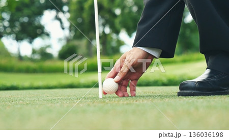 Businessman playing Golf ball practice hit swing on tee in golf club outdoor green field with recreation sport training course. Business men with black Suit driving golf Hobbies Businessman playing Golf ball practice hit swing on tee in golf club outdoor green field with recreation sport training course. Business men with black Suit driving golf Hobbies 136036198
