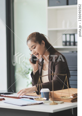 Confident Lawyer Working at Desk: A young Asian female lawyer confidently works on her laptop, surrounded by legal symbols. She exudes professionalism and competence in her modern office setting. 136040902