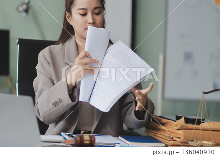 Confident Lawyer Working at Desk: A young Asian female lawyer confidently works on her laptop, surrounded by legal symbols. She exudes professionalism and competence in her modern office setting. 136040910
