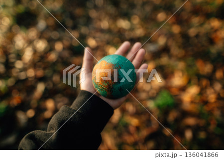 Wooden globe in hand, bokeh background of dried leaves, environmental conservation concept. 136041866