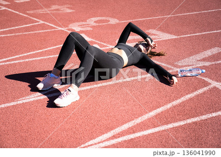 tired fit woman in sportswear resting after workout or running on a treadmill rubber stadium, holding a bottle of water, taking a break during training tired fit woman in sportswear resting after workout or running on a treadmill rubber stadium, holding a bottle of water, taking a break during training 136041990