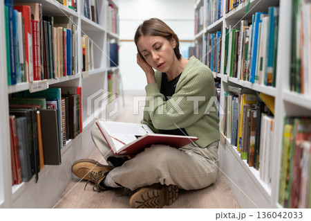 Pretty concentrated adult female sit on floor between bookshelves, reading interesting book on knees 136042033