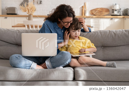 Mom embracing preschool kid playing games on tablet during break from remote work on laptop computer 136042047