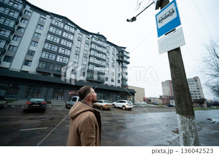 Man waiting at a bus stop while observing a nearby urban landscape on a cloudy day with modern buildings in the backdrop 136042523