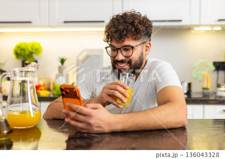 Arabian young adult guy drinking fresh orange juice in kitchen at home reading news on smartphone 136043328