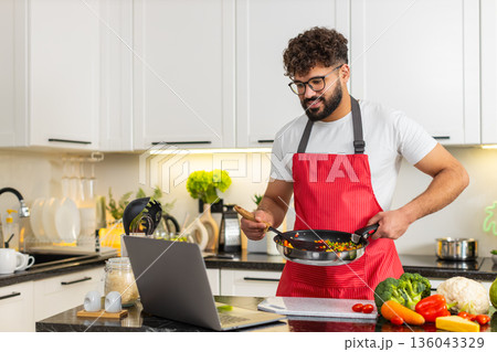 Arabian young adult man tossing hot vegetables in frying pan while watching laptop recipe in kitchen 136043329