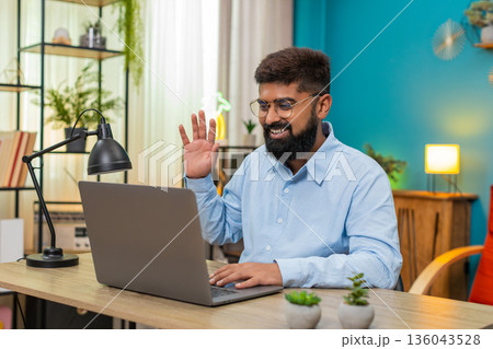 Indian man waves greeting speaks via laptop webcam in video call smiles during online conference web Indian man waves greeting speaks via laptop webcam in video call smiles during online conference web 136043528