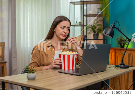 Young woman watching dramatic movie on laptop eating popcorn nervously reacting to intense scene 136043535