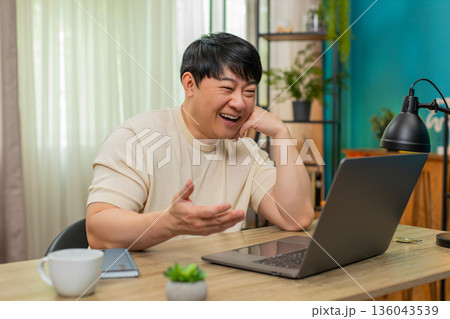 Asian man smiling watching nostalgic video on laptop, resting head on hand, enjoying warm memories Asian man smiling watching nostalgic video on laptop, resting head on hand, enjoying warm memories 136043539